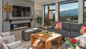 A modern living room with a flat-screen TV, fireplace, grey sofa, and large windows offering a mountain view.