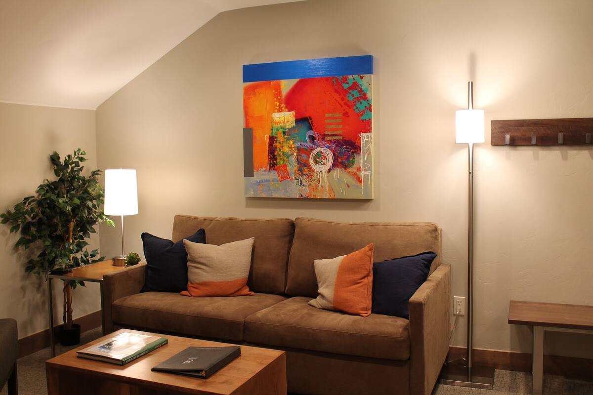 Ein modernes Wohnzimmer mit einem braunen Sofa, einem Couchtisch, einer Stehlampe und einem farbenfrohen abstrakten Gemälde an der Wand.