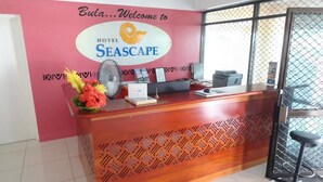Een hotel receptiebalie met een houten toonbank, een ventilator, een bloemenvaas en een bord met de tekst 'Bula...Welkom bij HOTEL SEASCAPE'.