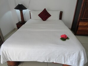 Una cama bien hecha con una colcha blanca, una sola almohada roja y una pequeña flor roja sobre la cama.
