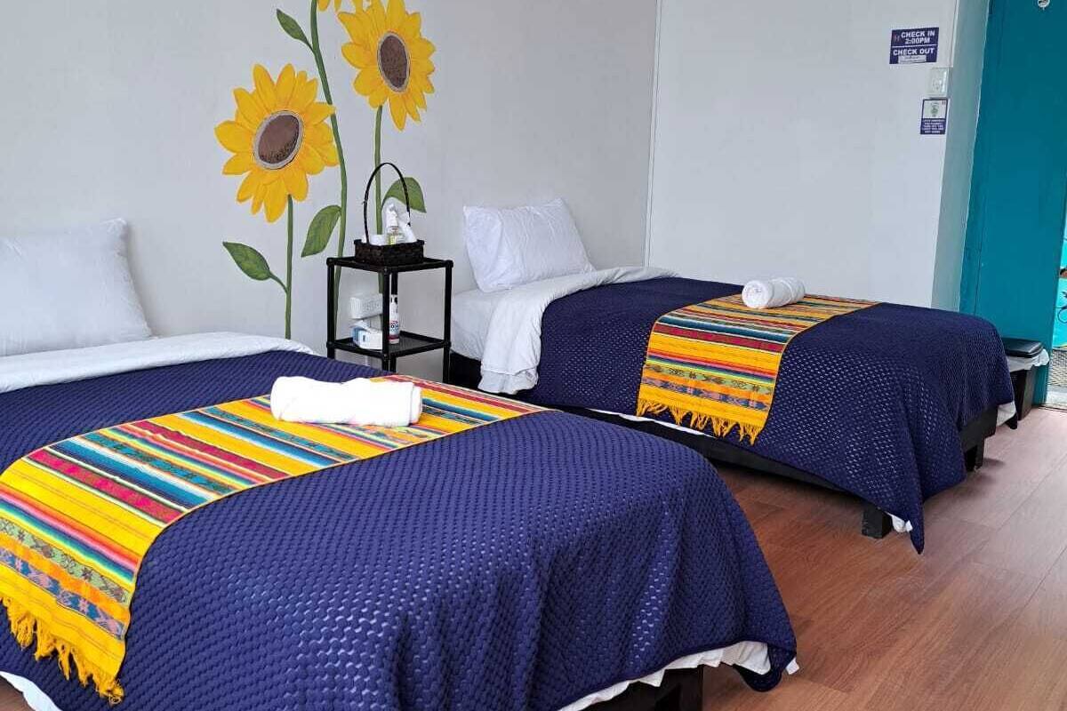 A hotel room with two beds, each with a blue bedspread and a colorful blanket. There is a small table with a white lamp and a decorative item.