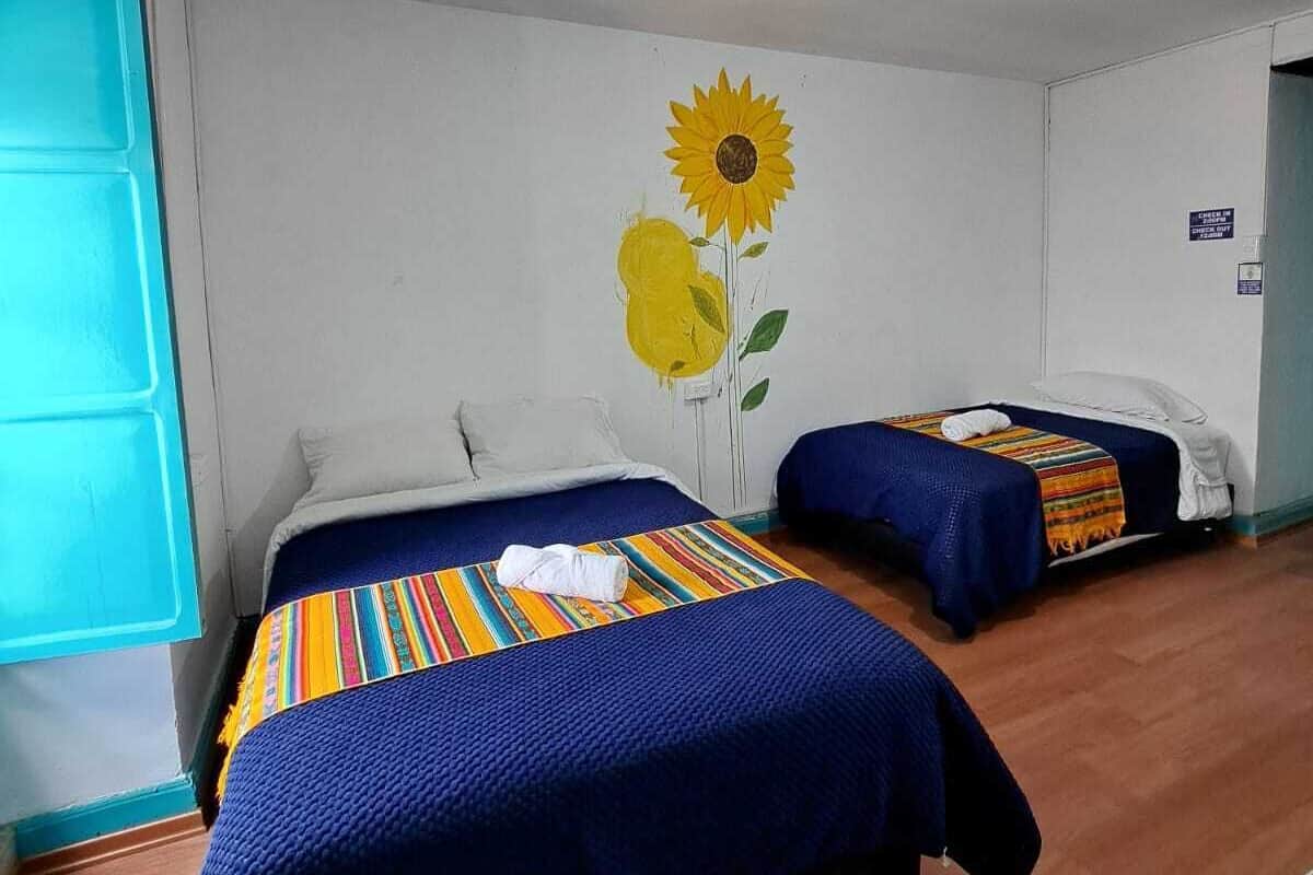 A room with two beds, each with a blue bedspread and a colorful striped blanket. There is a large sunflower mural on the wall.