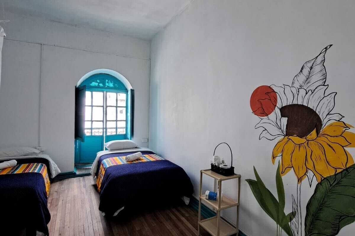 A room with two beds, a wooden floor, a window with a blue frame, and a wall mural of a sunflower.