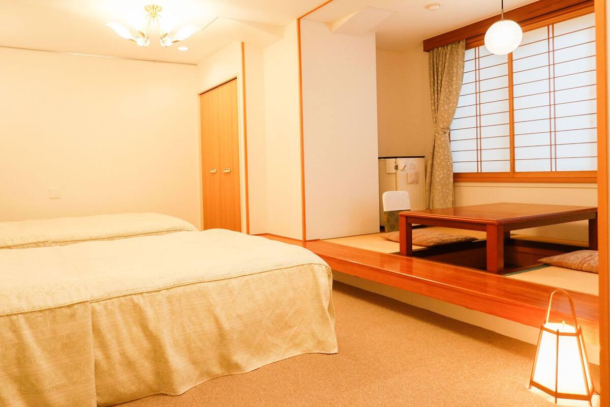 A bedroom with two beds, a wooden table, a lamp, and a window with a view.