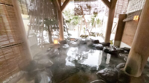 Un área de aguas termales con escalones de piedra que conducen a una piscina, persianas de bambú y una estructura de madera.