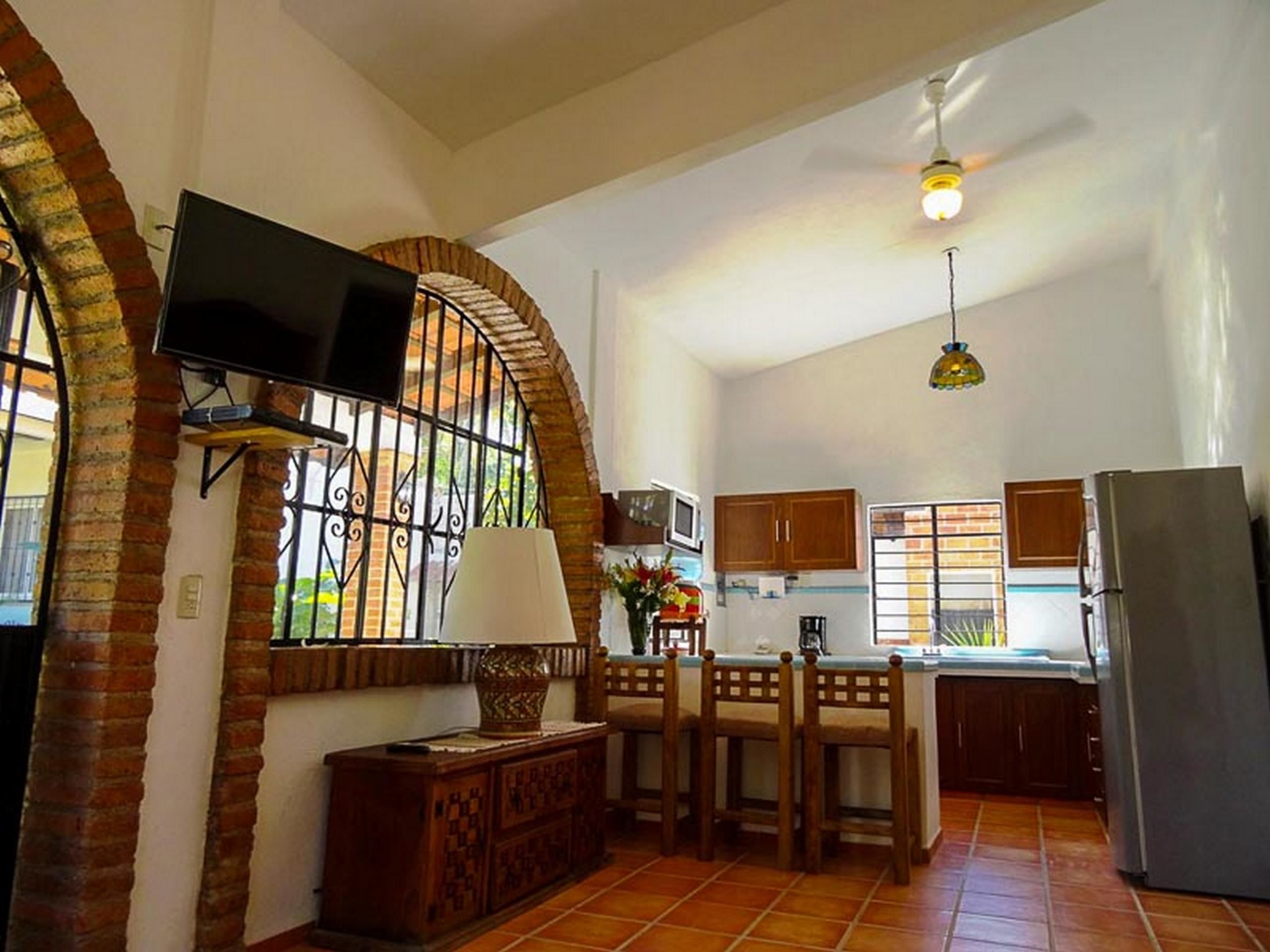 A spacious kitchen with a dining area, a TV mounted on a brick wall, and a ceiling fan.