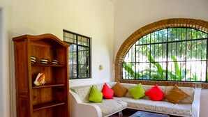 A living room with a wooden cabinet, a sofa with colorful cushions, a small table, and a large arched window with greenery outside.