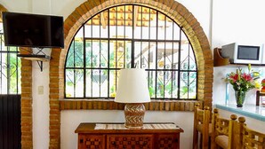 A room with a TV mounted on the wall, a wooden cabinet with a patterned top, a window with metal bars, and a chair with a wooden frame.