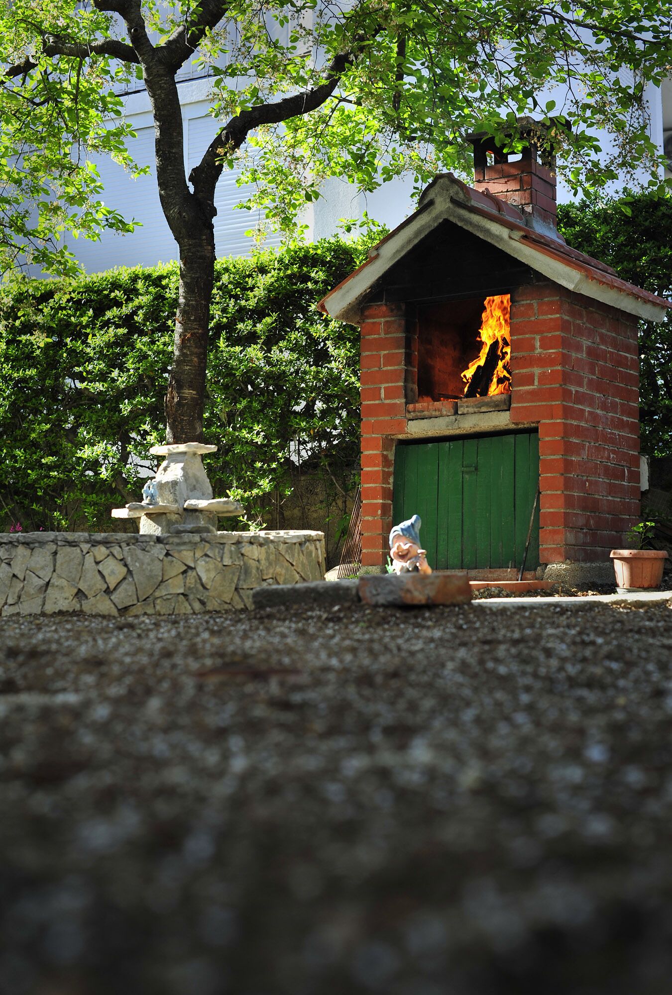 A brick outdoor oven with a green door, surrounded by a stone wall and a tree.