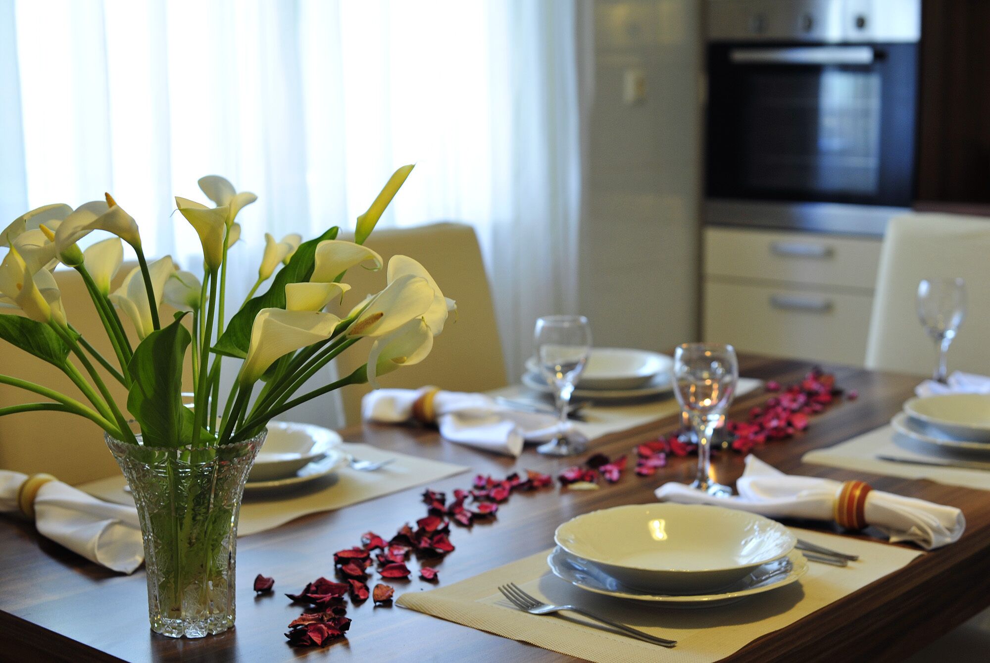 A dining table set for four with white plates, glasses, and a vase of white flowers.