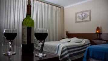 A hotel room with a bed, a bedside table with a bottle of wine and two glasses, a lamp, and a wall clock.
