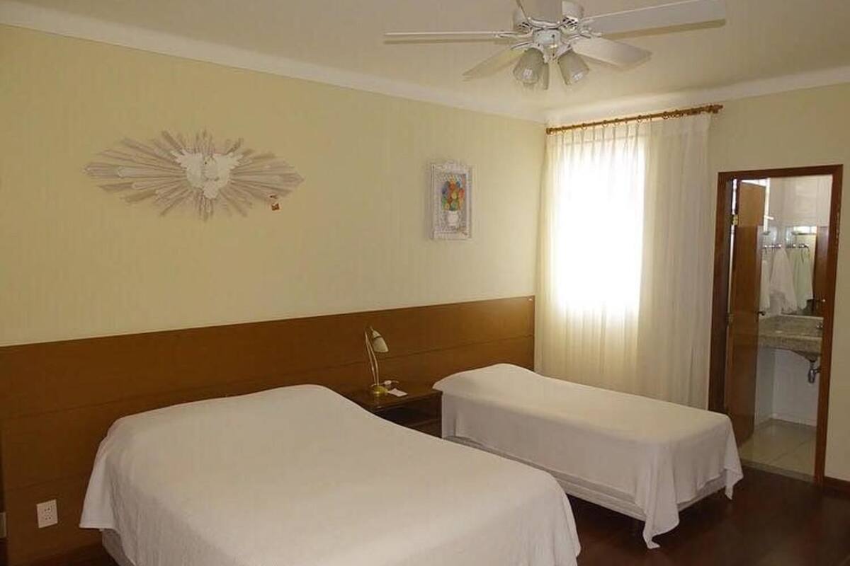 A hotel room with two single beds, a ceiling fan, a nightstand with a lamp, a decorative wall piece, a framed picture, and a bathroom visible through a door.