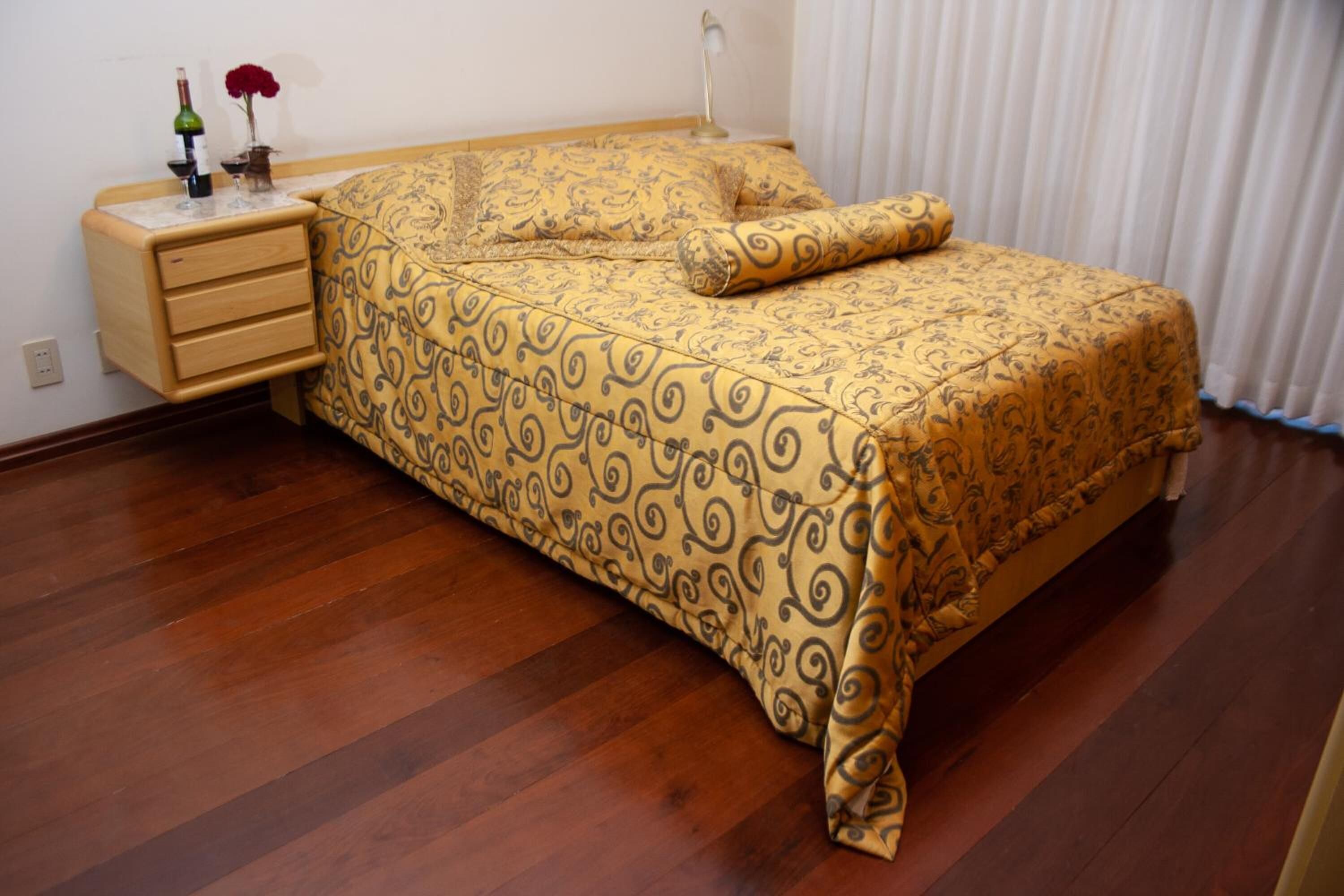 A bedroom with a patterned bedspread, wooden nightstand, and a bottle of wine.