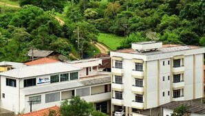 A building with a flat roof and multiple windows, surrounded by lush greenery and a hilly landscape. - Apart Hotel Água Limpa (Prados)