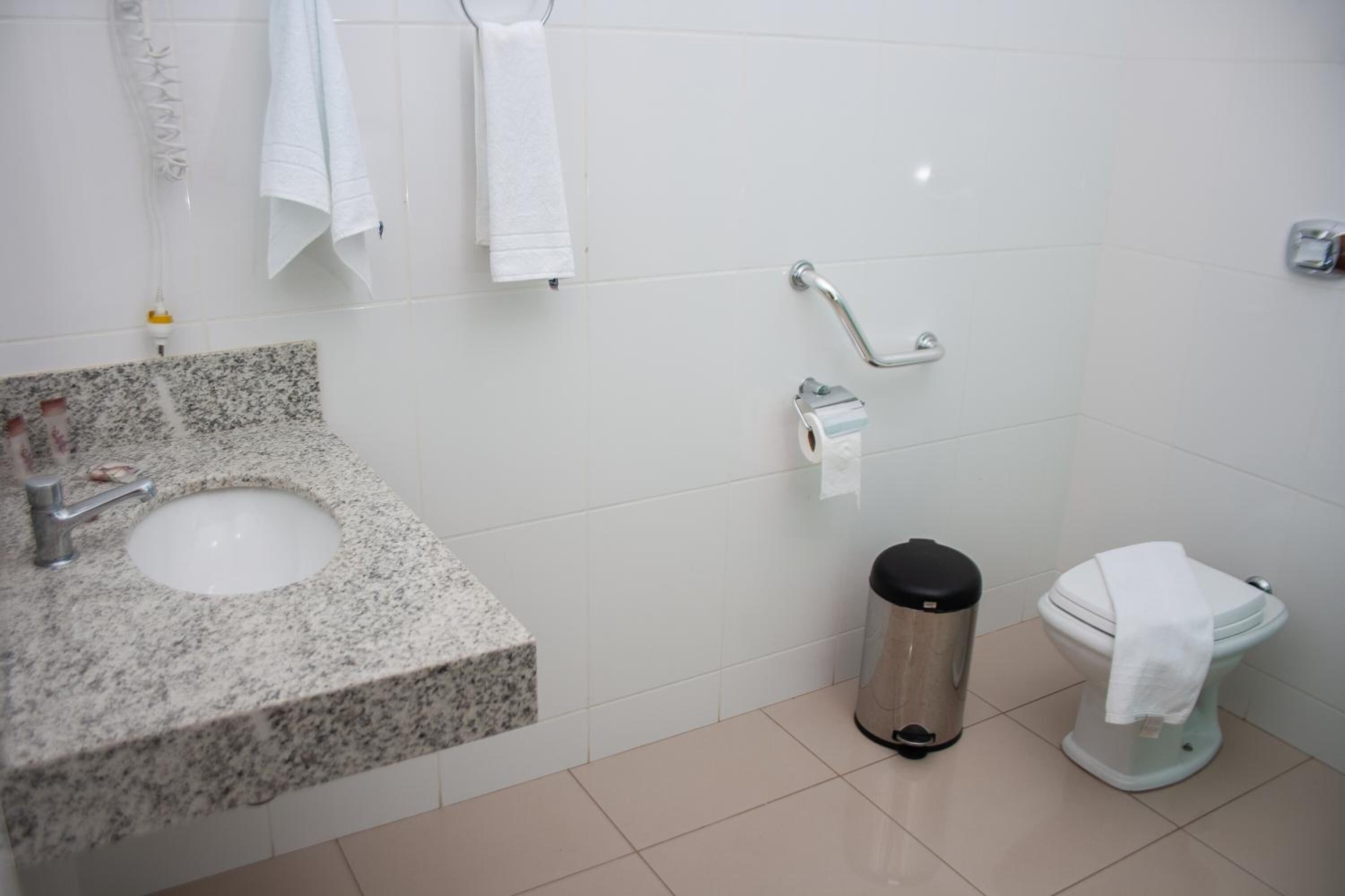 A bathroom with a granite sink, white tiles, a toilet, a trash can, and a handrail.