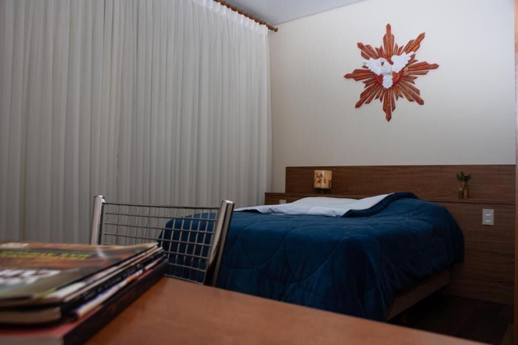 A hotel room with a bed, a headboard, a wall decoration, a window with curtains, and a table with books.