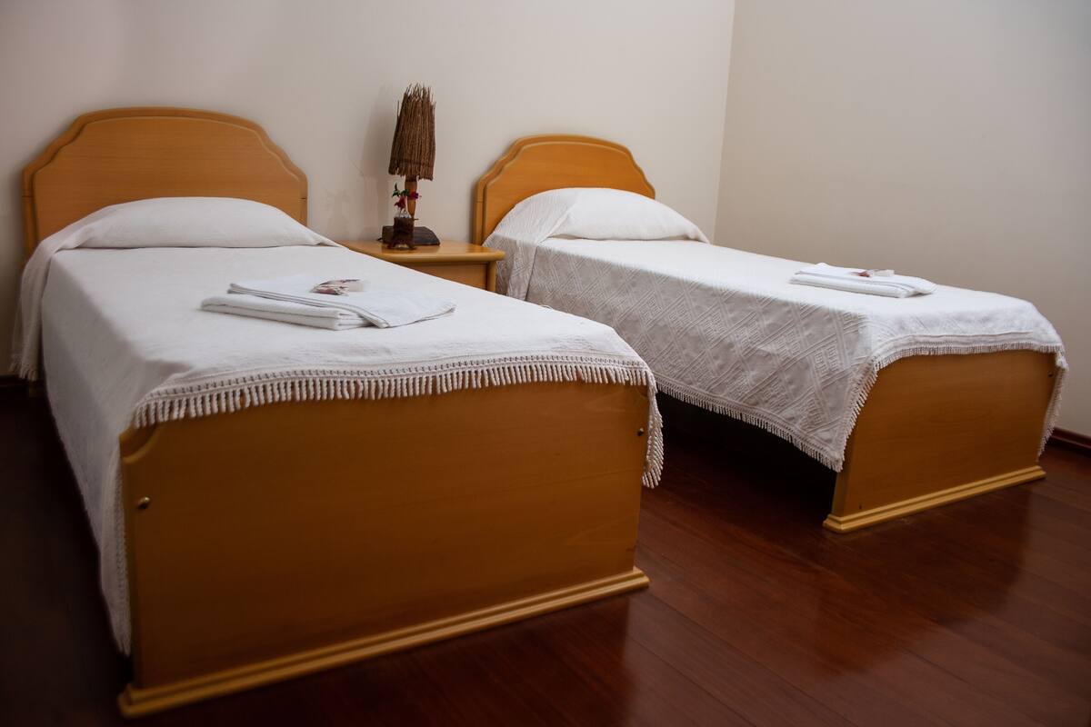 Two single beds with wooden frames and white bedding, a small bedside table with a lamp, and a plain wall.