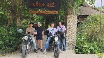 A group of people posing with motorcycles in front of a rustic stone building with a sign reading "ĐƠNG QUÊ Homestay & Restaurant".