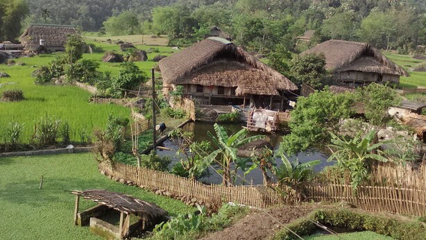 A rural village with thatched-roof houses, a bamboo fence, and a water body. - Dong Que Homestay-Tours-Motorbike, Car rental, Hiking (Ha Giang 1)