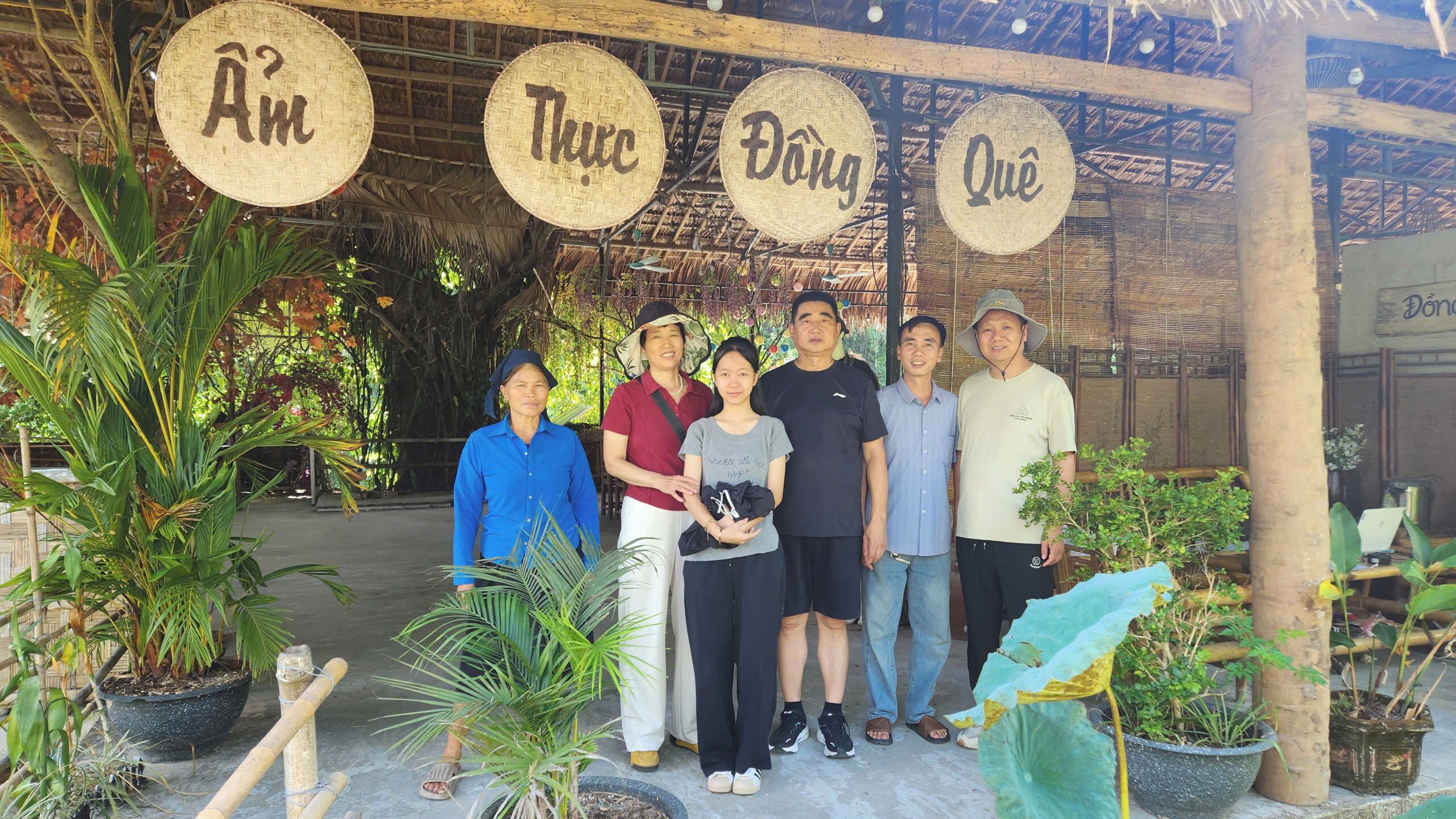 A group of people standing under a thatched roof structure with woven signs displaying names.