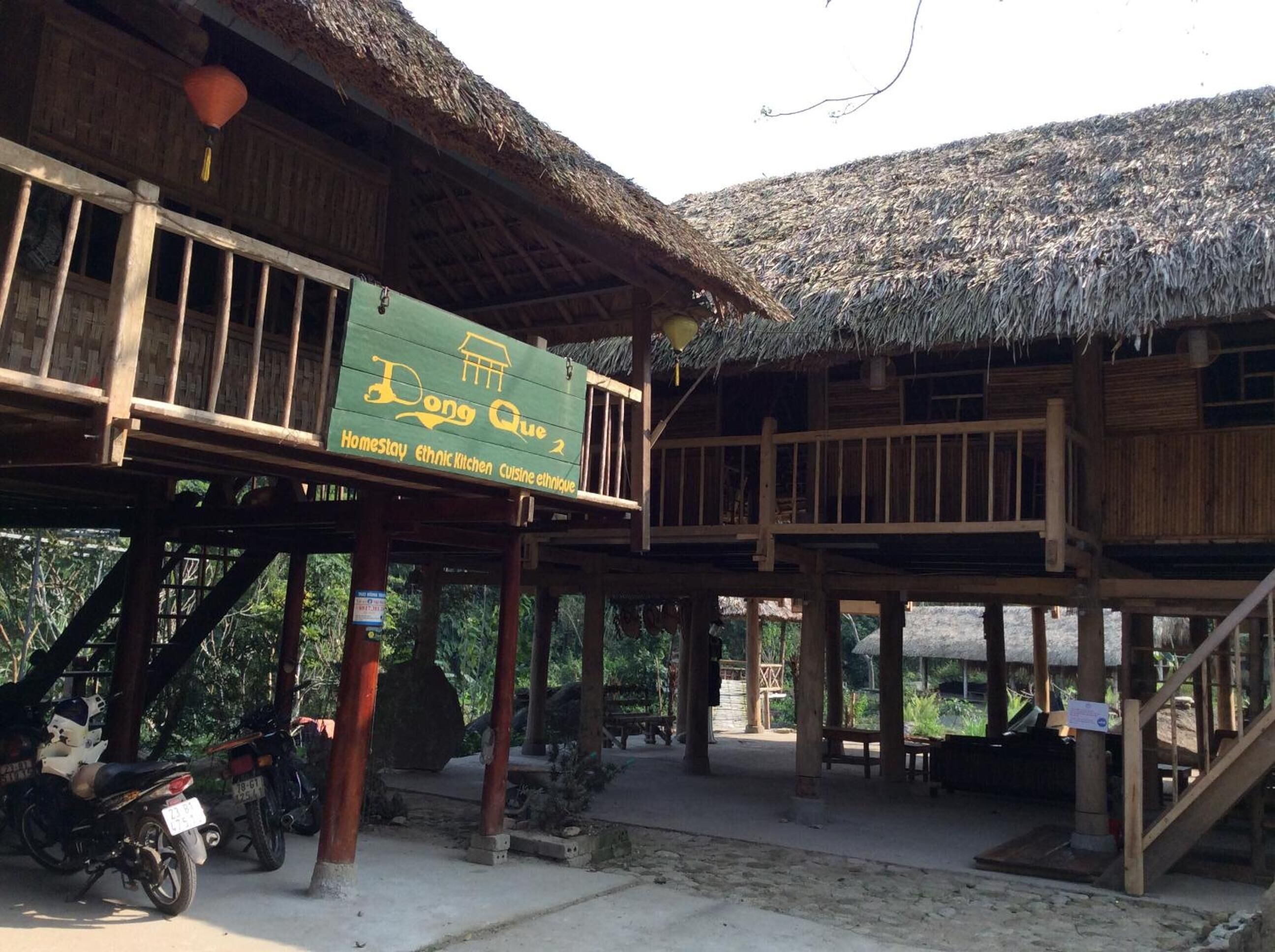 A wooden structure with a thatched roof, a signboard advertising 'Dong Que 2 Homestay ethnic kitchen cuisine ethnic', and a staircase leading to an upper level.