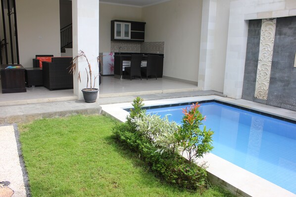A swimming pool with a grassy area, a small garden, and a kitchen area with a dining set.