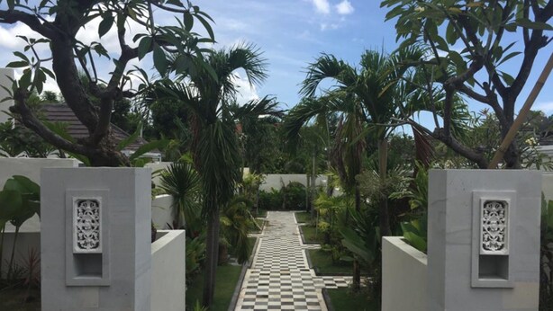 Un sendero que atraviesa un jardín con palmeras y un suelo a cuadros, bordeado por paredes blancas con paneles decorativos.