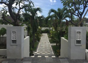 Um caminho serpenteia por um jardim com palmeiras e piso xadrez, ladeado por paredes brancas com painéis decorativos.