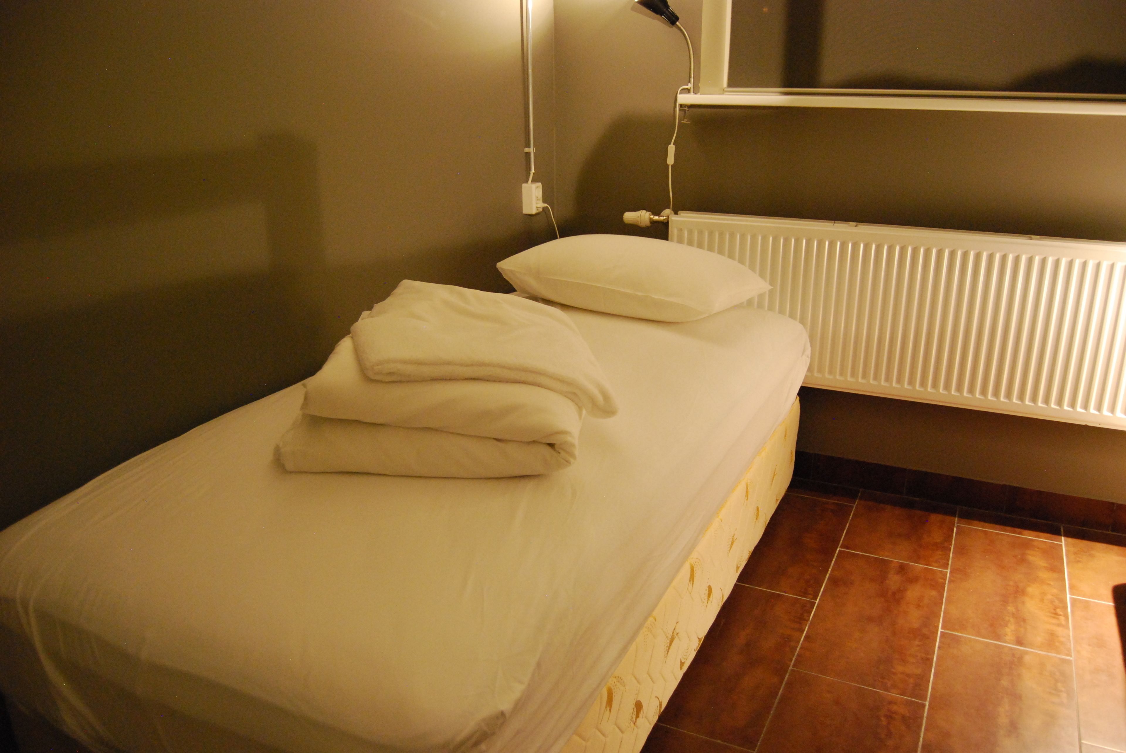 Un lit simple avec du linge de lit blanc et des oreillers, un radiateur blanc et un sol carrelé.