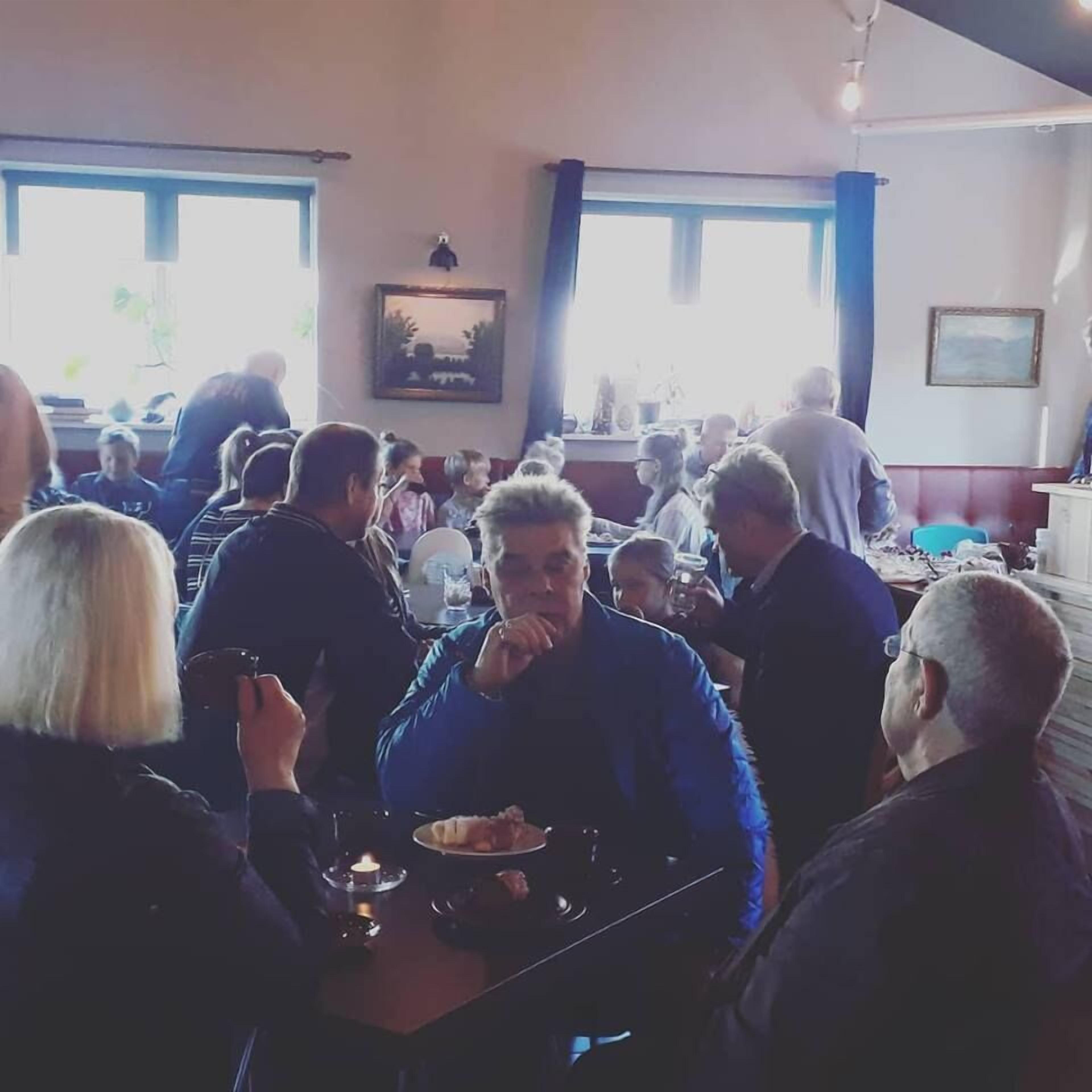 A crowded restaurant with tables and chairs, a painting on the wall, and windows with curtains.