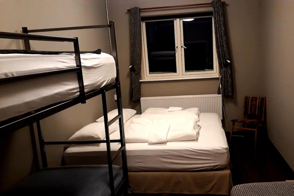 A dormitory room with bunk beds, a single bed, a window with curtains, a radiator, and a chair.