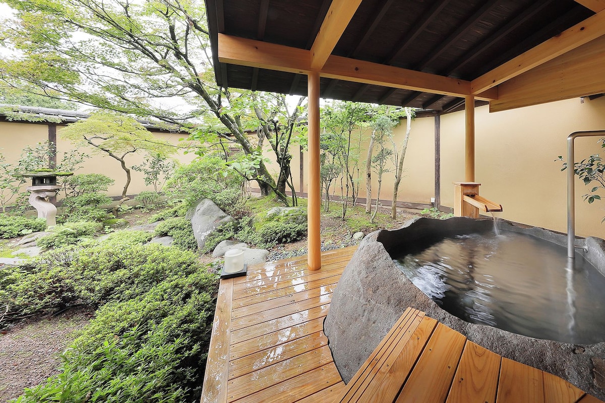 A hot spring area with a wooden deck, a stone hot tub, and surrounding greenery.