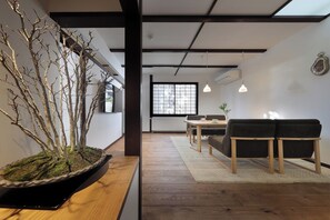 Ein modernes Interieur mit Holzboden, einer Essecke mit Tisch und Stühlen, einer Couch, einer Zimmerpflanze und einem großen Fenster mit Jalousien.
