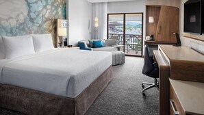 Ein Hotelzimmer mit einem großen Bett, einem Schreibtisch, einem Sessel, einem Fernseher und einem Balkon mit Blick auf ein Stadion.