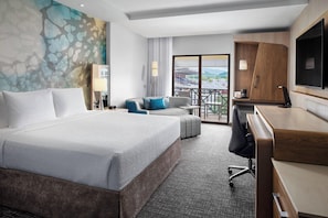 Ein Hotelzimmer mit einem großen Bett, einem Schreibtisch, einem Sessel, einem Fernseher und einem Balkon mit Blick auf ein Stadion.