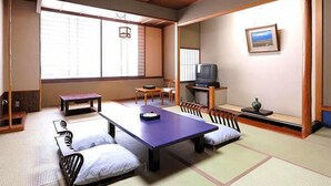 Ein traditioneller japanischer Raum mit Tatami-Boden, einem niedrigen Tisch, Kissen, einem Fernseher und einem gerahmten Bild an der Wand.