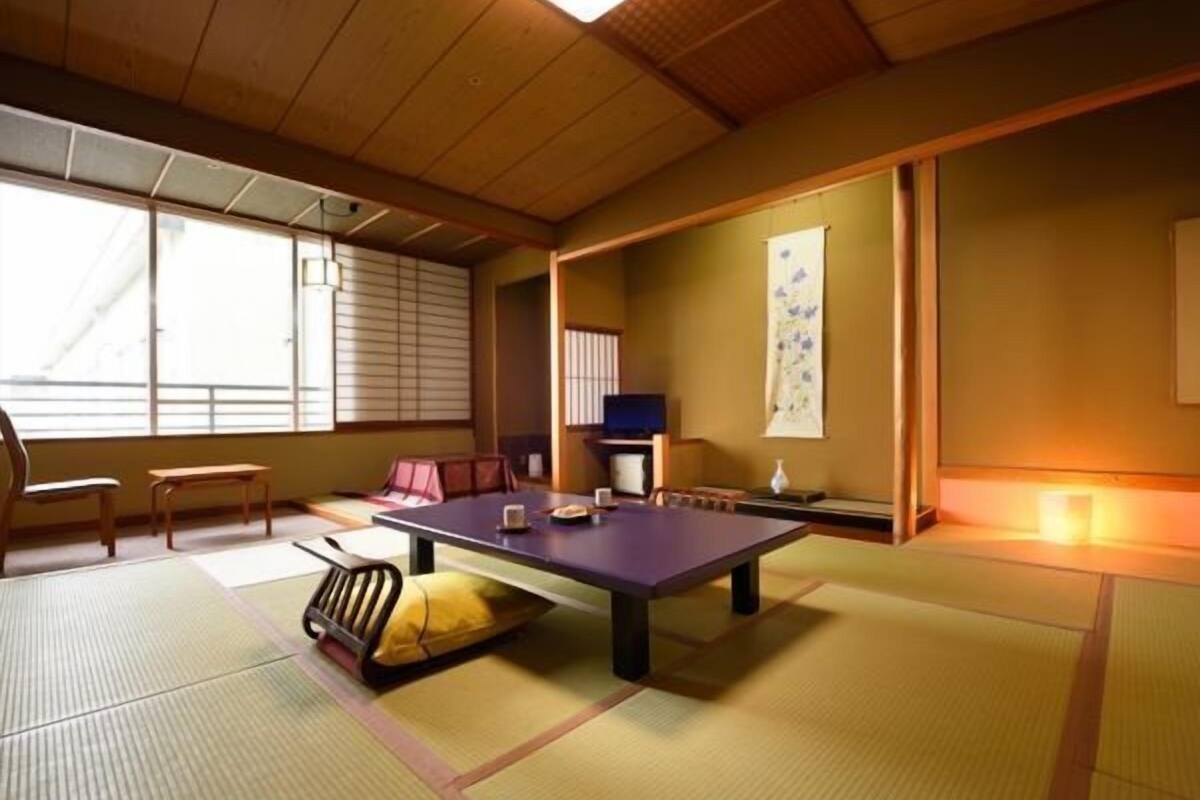 Une pièce avec un sol recouvert de tatamis, une petite table, des chaises, un téléviseur et une porte coulissante.