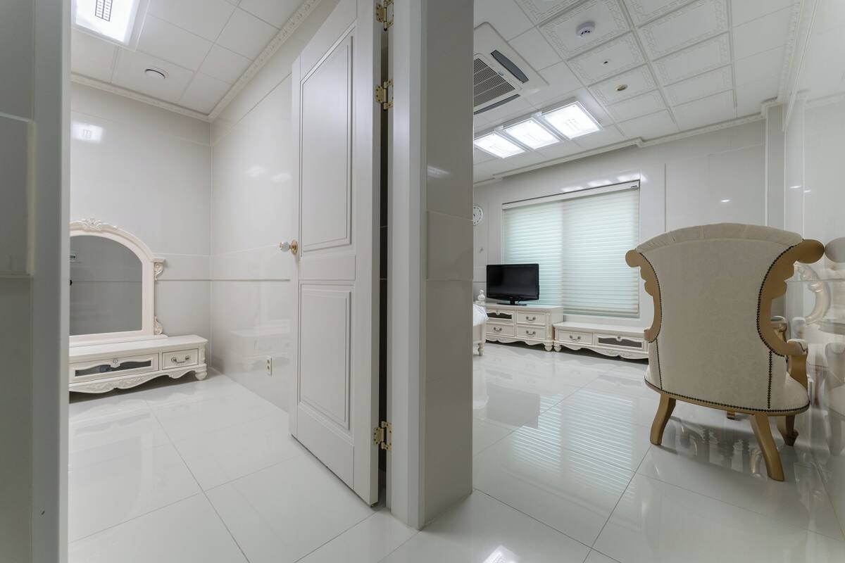 Une pièce avec un sol carrelé de carreaux blancs, une grande porte blanche, une coiffeuse avec un miroir, un téléviseur posé sur un meuble blanc et un fauteuil beige recouvert de tissu.
