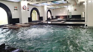 Un área de aguas termales con una amplia piscina interior, varios ventanales y una pantalla digital en el techo.