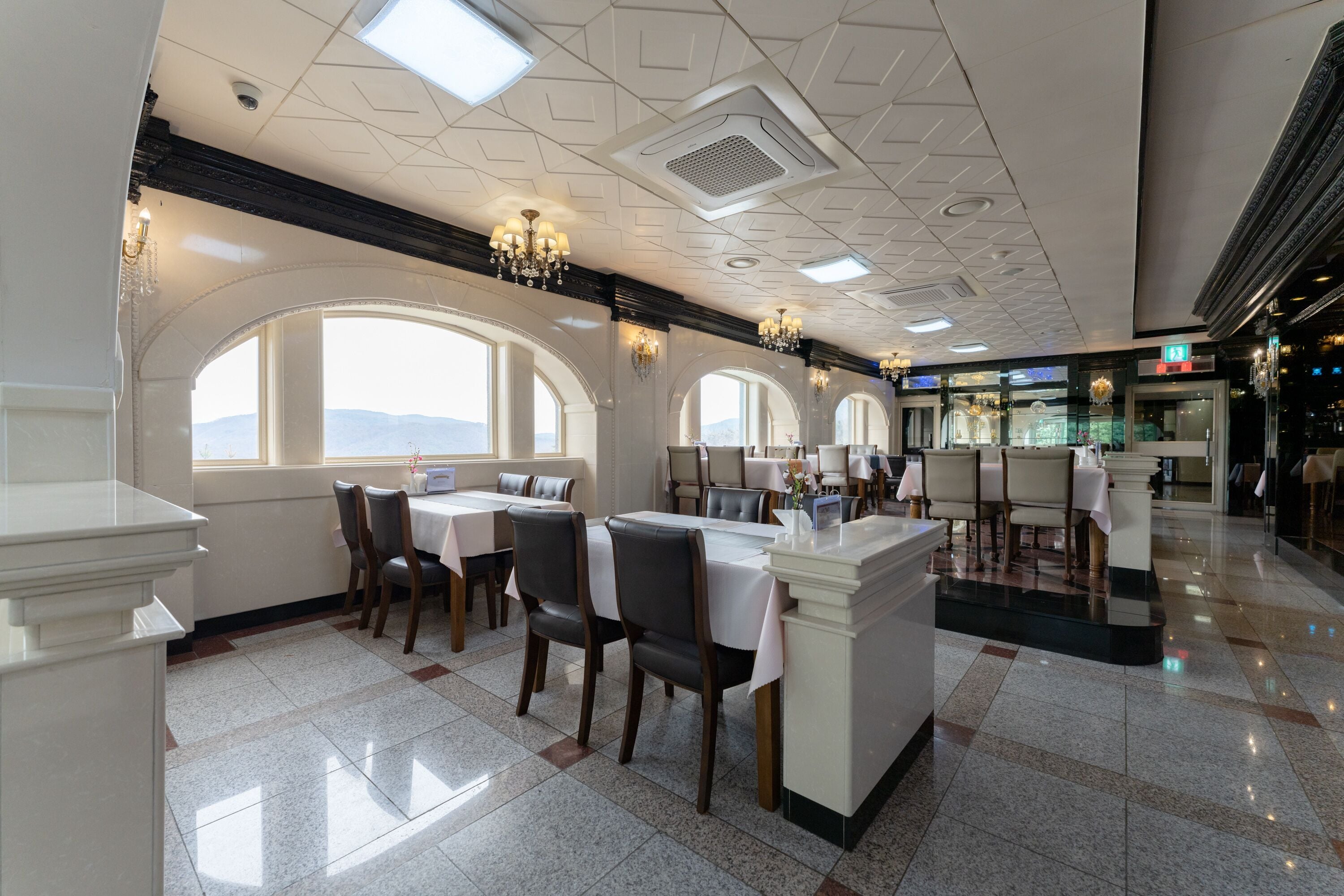 A restaurant with a high ceiling, chandeliers, and large windows offering a view. Tables are arranged around a central island.
