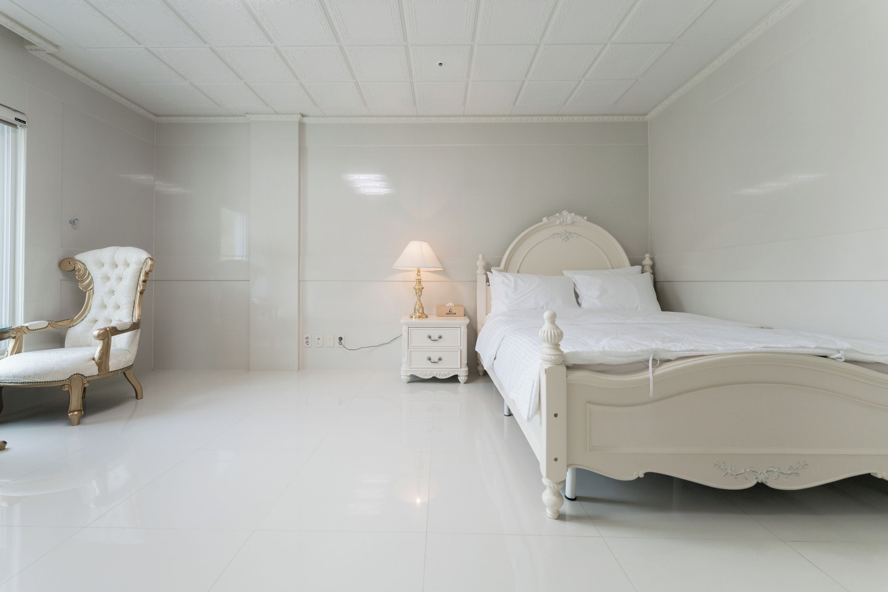 Un dormitorio con cama blanca, mesita de noche, lámpara y silla.