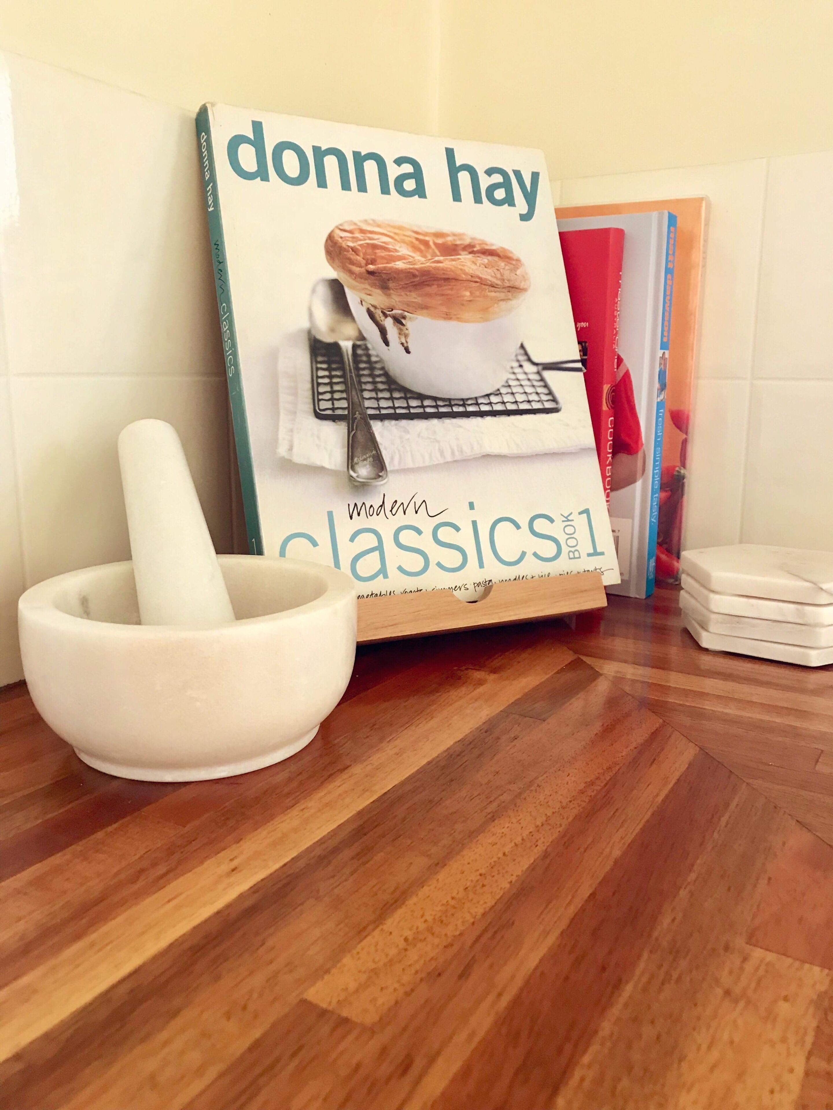 Eine Küche mit einem Regal, auf dem das Buch 'donna hay classics' steht, einem weißen Mörser und Stößel sowie einem Holzboden.