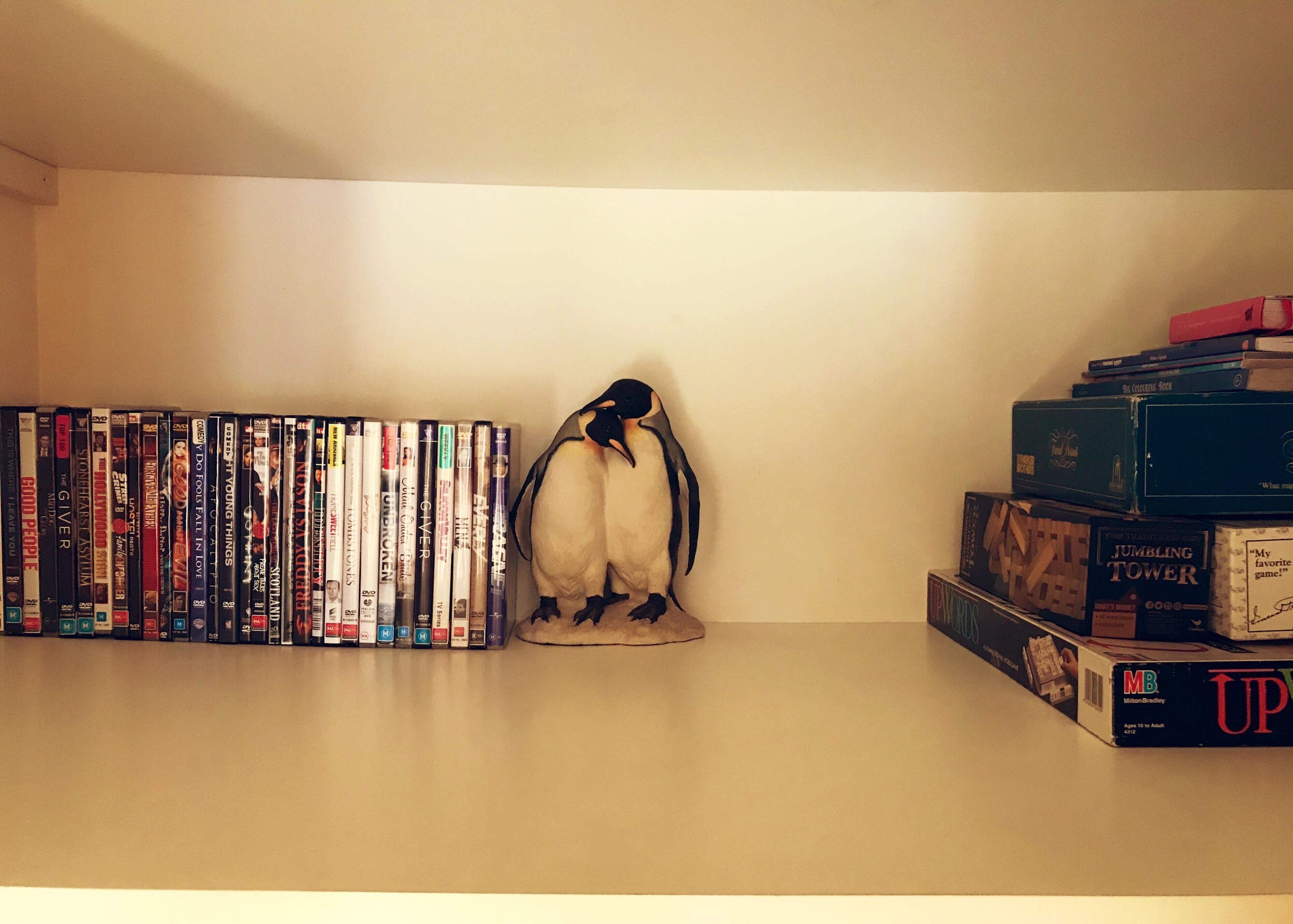A shelf with a collection of DVDs and books, a penguin figurine, and a stack of board games.
