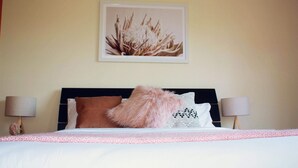 Una cama bien hecha con un almohadón rosa esponjoso y otro blanco, un cuadro en la pared y dos lámparas de noche.