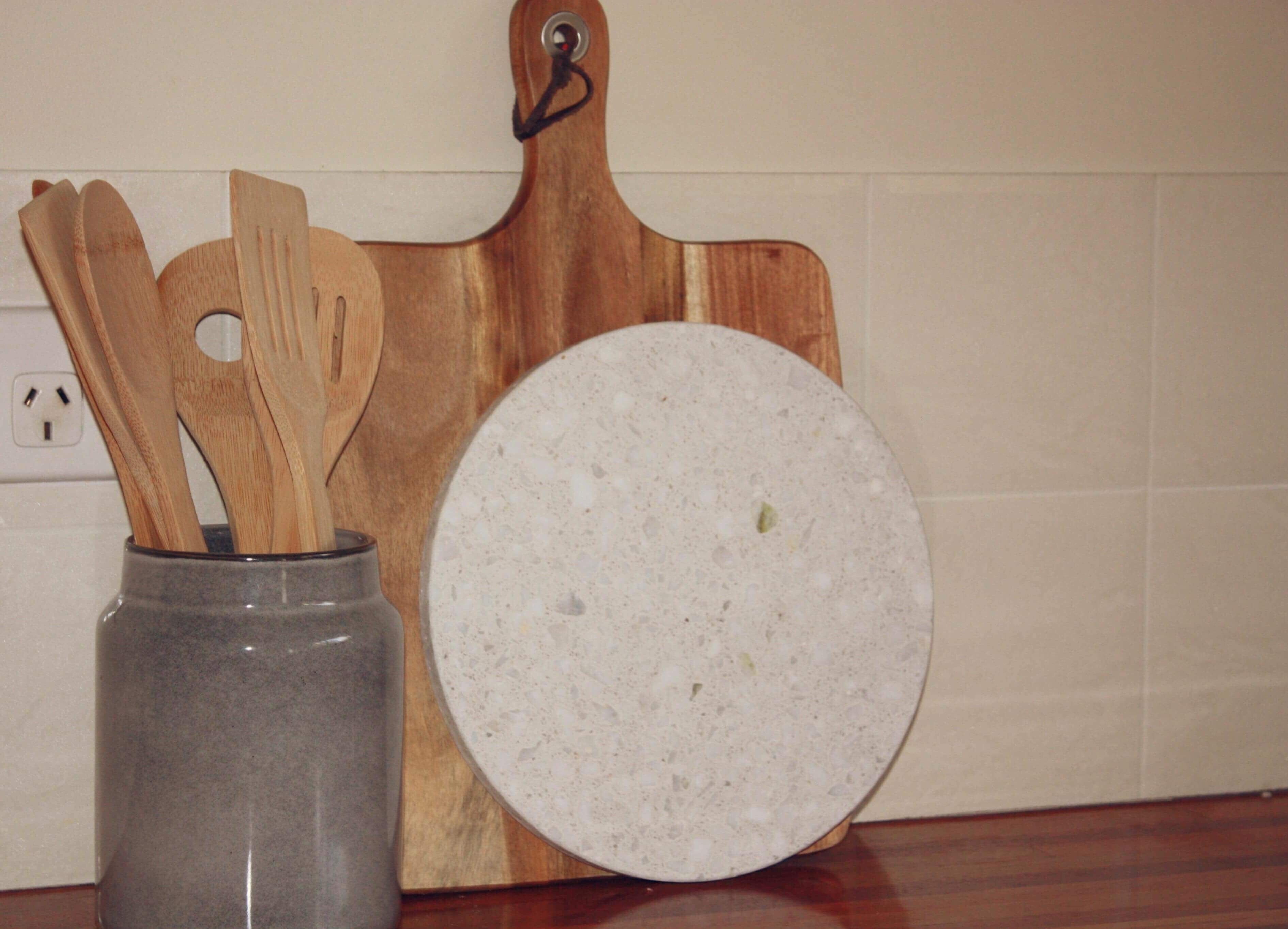 A kitchen counter with a wooden cutting board, a round white plate, and a gray vase holding wooden utensils.
