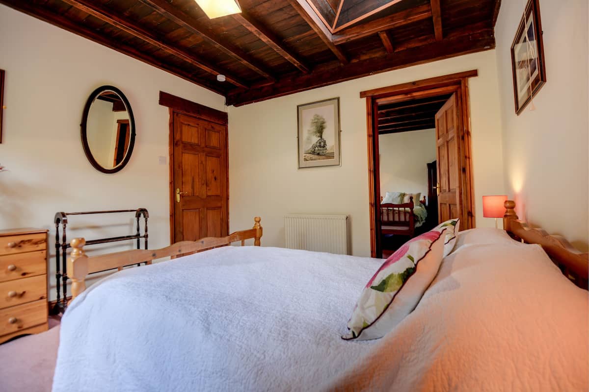 Un dormitorio con una cama grande, cabecera de madera, cómoda de madera, un espejo y una puerta que da a otra habitación.
