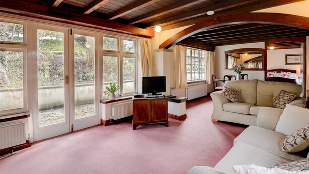 Ein geräumiges Wohnzimmer mit Holzboden, großen Fenstern, einem beigefarbenen Sofa und einem Fernseher auf einer Holzkommode.