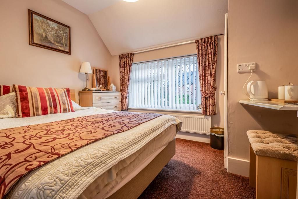 A bedroom with a bed, a nightstand, a lamp, a window with curtains, a radiator, and a small table with a kettle.
