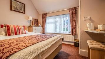 A bedroom with a bed, a nightstand, a lamp, a window with curtains, a radiator, and a small table with a kettle.