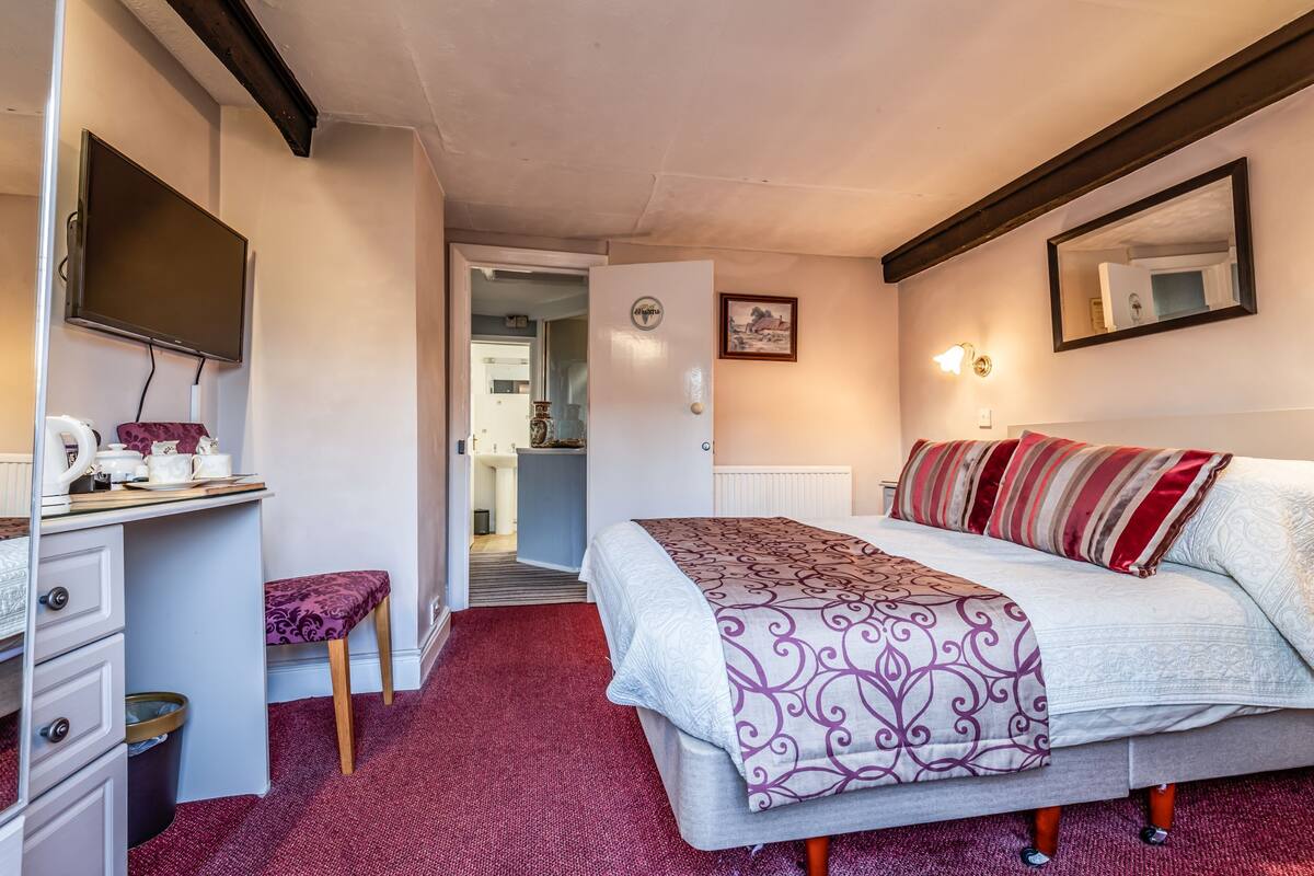 A bedroom with a bed, a desk with a chair, a television, and a door leading to another room.